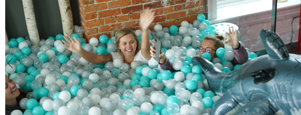 Two people are submerged in a pool of blue and white plastic balls, playfully throwing them into the air. A brick wall and a window are visible in the background, and there's an inflatable dolphin toy among the balls.