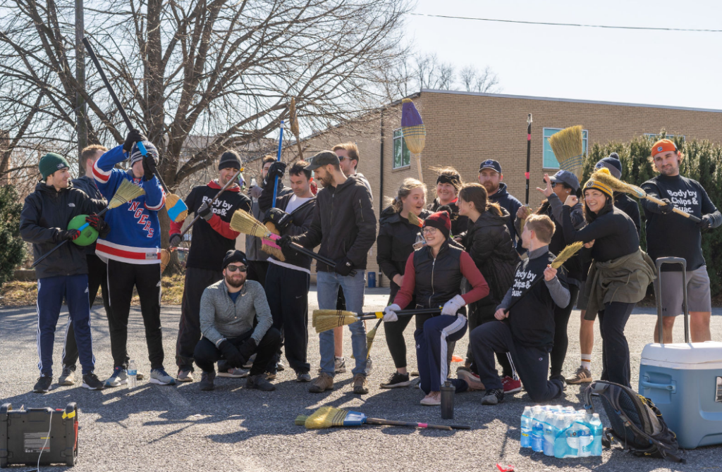 A group of cheerful people posing with brooms outdoors, some wearing matching 'Bobby D's Brooms & Dance' t-shirts, suggesting a playful event or activity.