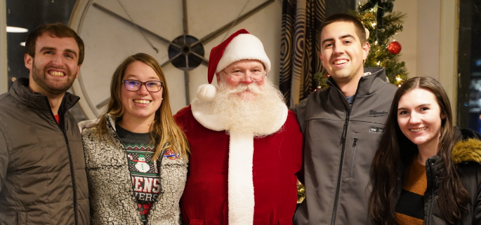 Four people posing with a person dressed as Santa Claus indoors with a Christmas tree and festive decorations in the background.