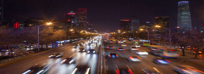 A busy city street at night with streaks of light from moving vehicles, surrounded by illuminated buildings under a dark sky.