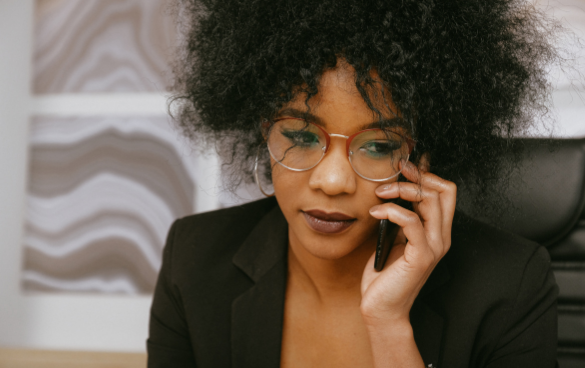 A woman with curly hair wearing glasses and a black blazer is speaking on a mobile phone, looking focused and serious.