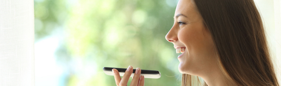 Side profile of a smiling woman speaking into a smartphone, with a bright window in the background.