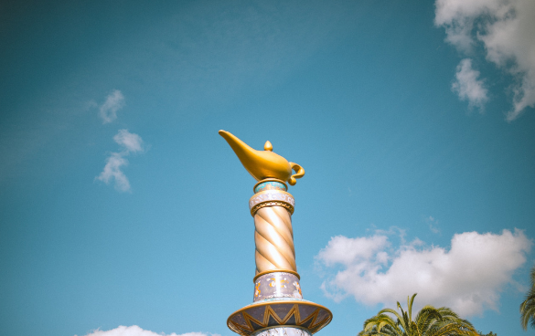 A stylized golden lamp on top of a decorative column against a blue sky with palm fronds at the bottom.