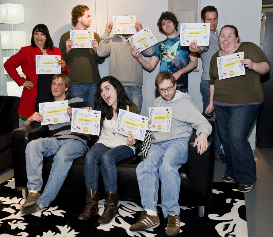 A group of eight people posing with humorous certificates, some standing and some seated on a black couch, in a room with modern decor, expressing a mix of excitement and casualness.