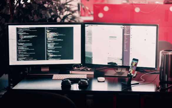 A dual-monitor setup on a desk with the left screen showing code and the right screen displaying a web browser. A keyboard, mouse, headphones, and a smartphone are also on the desk, with plants and a metallic container in the background.