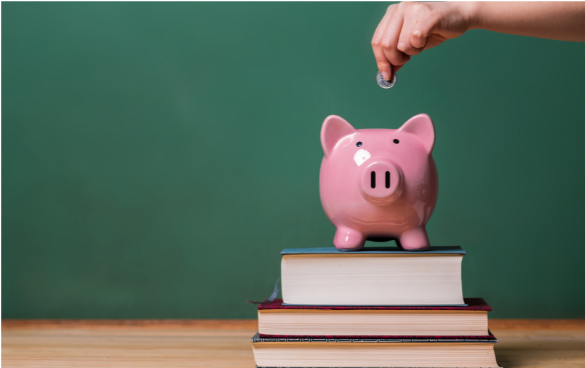 A person's hand is depositing a coin into a pink piggy bank on top of a stack of books with a green chalkboard in the background.