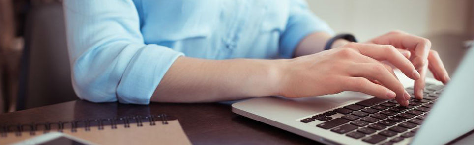 Person in a blue shirt typing on a laptop next to a spiral notebook on a desk.