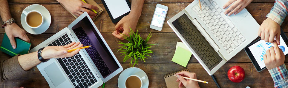 A top-down view of a collaborative workspace with multiple people's hands, two laptops, a tablet, notebooks, pens, coffee cups, a plant, sticky notes, and an apple on a wooden table.