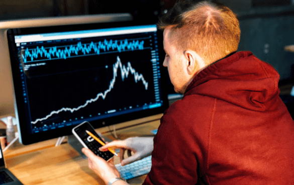 Person in red hoodie analyzing a fluctuating stock chart on a computer monitor while holding a smartphone.