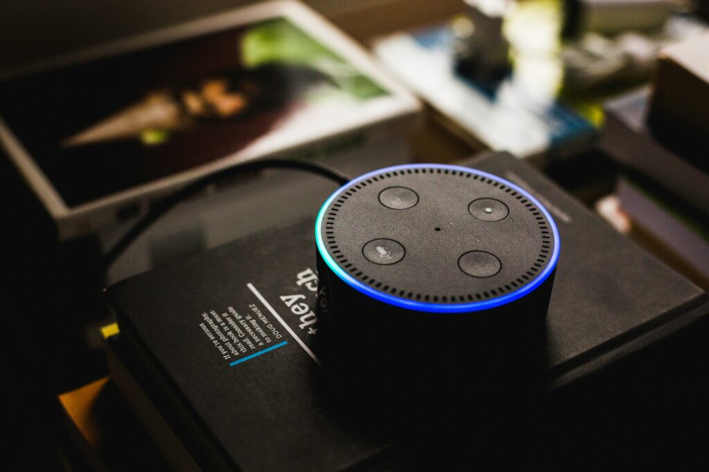 A smart speaker with an illuminated blue light ring is placed on a black book or box, with blurred items in the background.