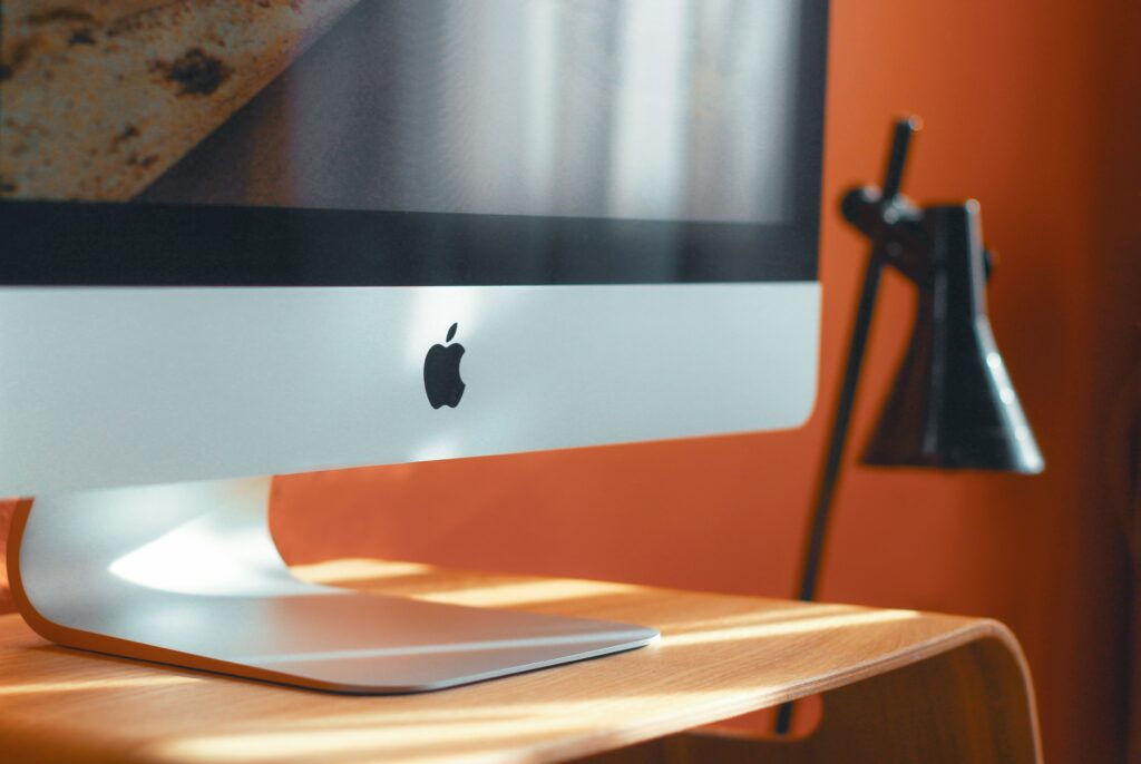 An Apple iMac on a wooden desk with a black desk lamp in the background and an orange wall.