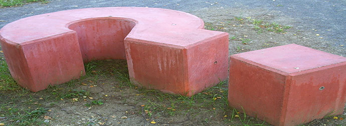 Red concrete outdoor furniture with a semi-circular bench and a separate square stool on a grassy area with a paved surface in the background.