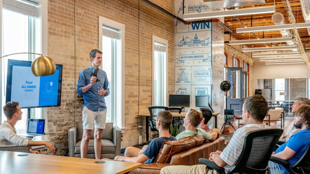 A man presenting to a group of seated individuals in a modern office space with a presentation screen displaying 'Proof ALL HANDS' dated '4/24/2019'. The setting is casual with a relaxed dress code and motivational office decor.