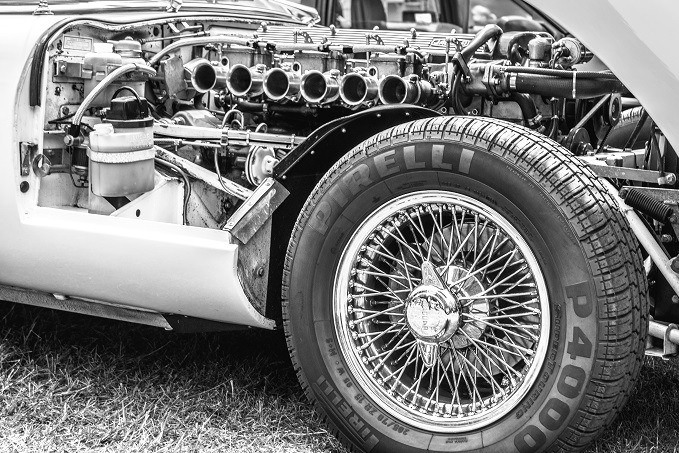 Close-up monochrome image of a vintage car's exposed engine and wire-spoke front wheel with a Pirelli P4000 tire.