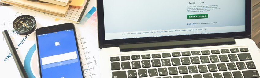 A workspace with a laptop open to a sign-up page, a smartphone showing the Facebook login screen, a compass, a pencil, and printed reports on a desk.