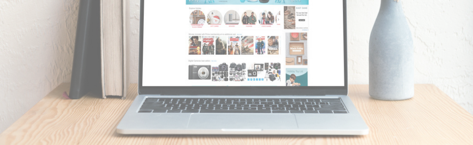 An open laptop on a wooden desk displaying a shopping website, flanked by two stacked books on the left and a ceramic vase on the right, against a white wall.