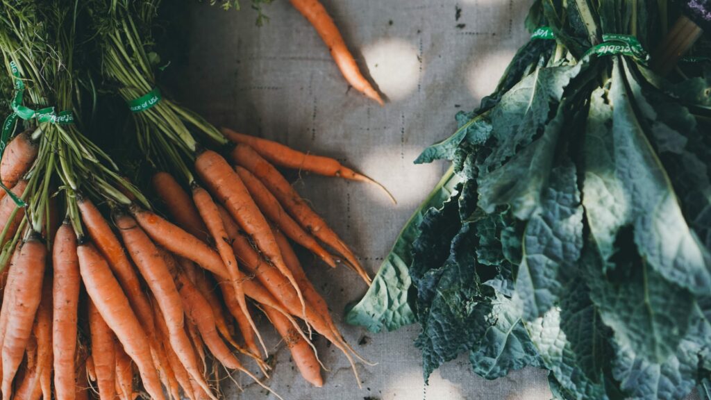 Freshly harvested bunch of carrots with green tops on the left and a bunch of curly kale on the right, both tied with green bands, on a textured surface.