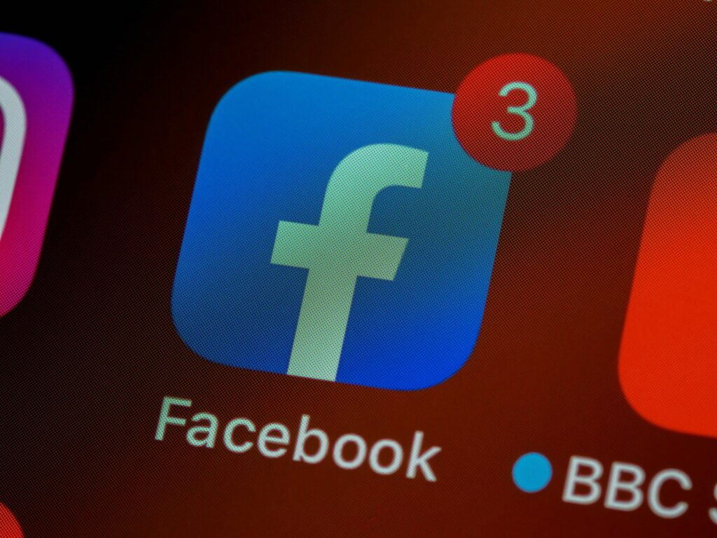 Close-up of the Facebook app icon with a red notification badge showing three new notifications on a smartphone screen, with other app icons partially visible in the background.