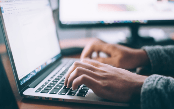 Person typing on a laptop with a second monitor in the background, indicating a dual-screen workspace.