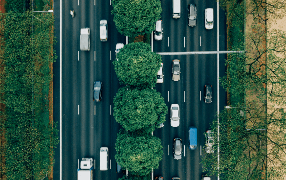 Aerial view of a multi-lane road with cars and trucks, bordered by lush trees on one side and a grassy area on the other.