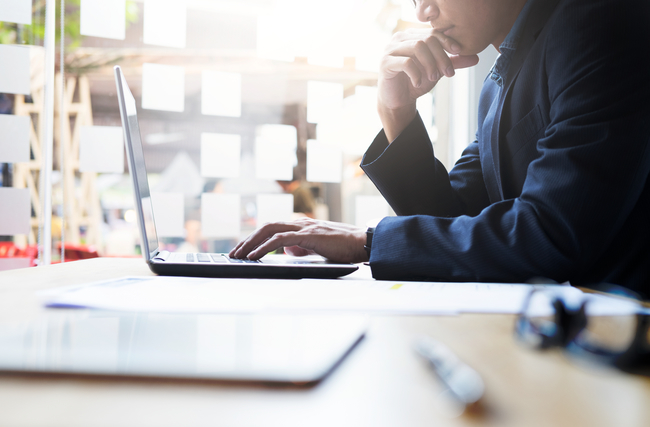 Person in deep thought working on a laptop at a desk with papers and glasses, in a bright office or cafe setting.