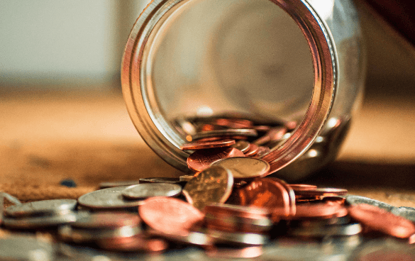 A glass jar tipped over with coins spilling out onto a wooden surface.