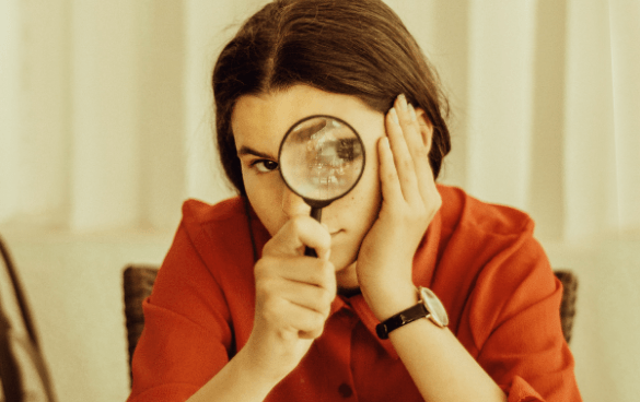 Person in a red shirt looking through a magnifying glass, which enlarges their right eye, while resting their head on their hand.