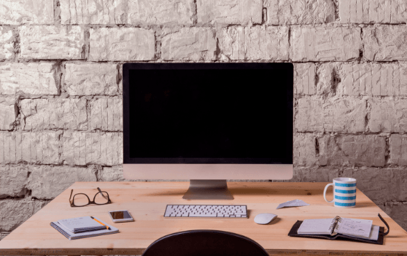 A tidy workspace with a computer monitor, keyboard, mouse, eyeglasses, smartphone, notepad, pen, clipboard, and a blue-striped mug on a wooden desk against a white brick wall.