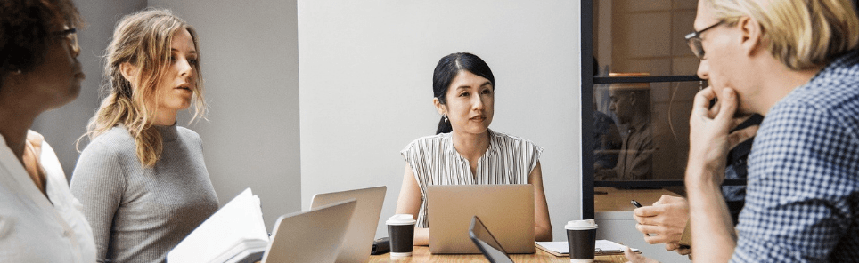 Four professionals engaged in a meeting with laptops and notebooks on the table, along with coffee cups, in a casual office environment.