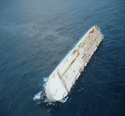 A large capsized ship with its red and white hull exposed, floating in a calm ocean.