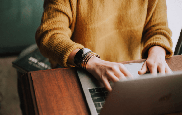 Person in a yellow sweater typing on a laptop at a wooden desk with bracelets on their wrist and books beside the laptop.