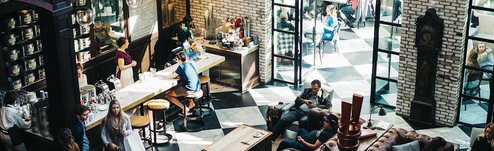 A lively café interior with patrons seated at tables, some using laptops, others conversing, and baristas working at the counter. The décor includes exposed brick walls, large windows, and a tall grandfather clock.