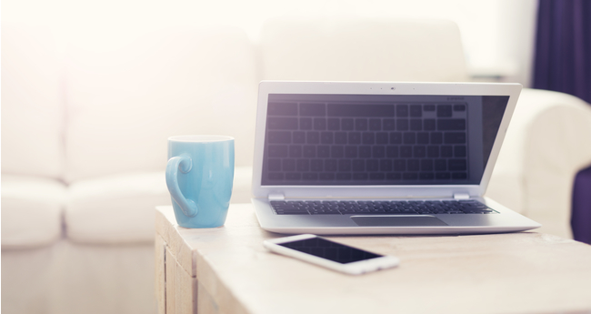 An open laptop, blue mug, and smartphone on a wooden table with a white couch in the softly lit background, indicating a home setting.