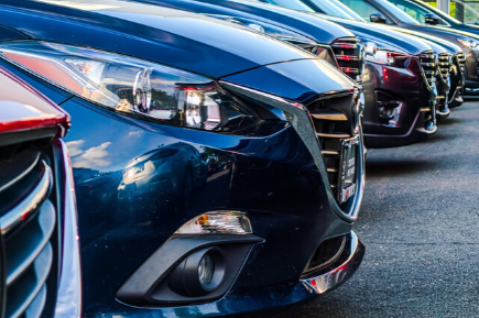 A row of parked cars at a dealership, focusing on the front left side of a dark-colored car with shiny exteriors and visible headlight and grille designs.