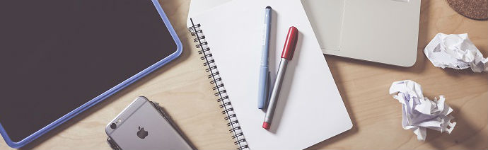 A workspace with a tablet, iPhone, closed laptop, notebook with pens, and crumpled paper on a wooden desk.