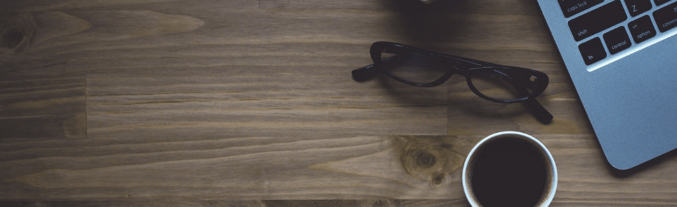 A wooden desk with a laptop, a pair of black eyeglasses, and a cup of coffee.