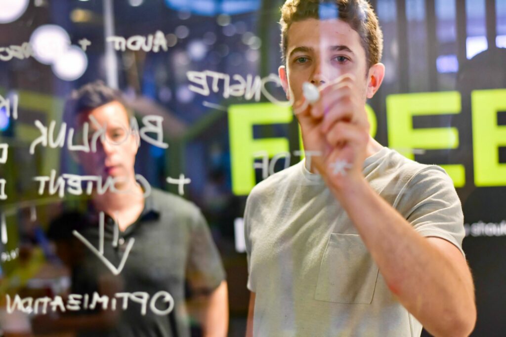 A young man writing mathematical equations on a transparent board with a marker, with another person observing in the background.