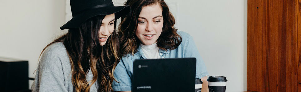 Two women, one wearing a black hat, looking at a laptop screen with a coffee cup beside the laptop in an indoor setting.