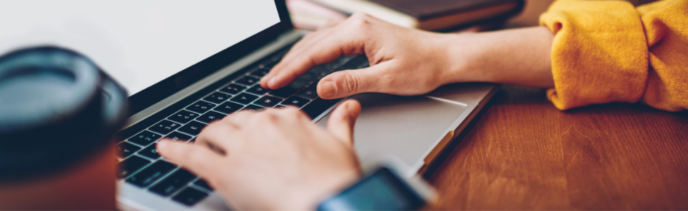 Close-up of two hands typing on a laptop keyboard with a person wearing a yellow sleeve and a watch in the foreground.