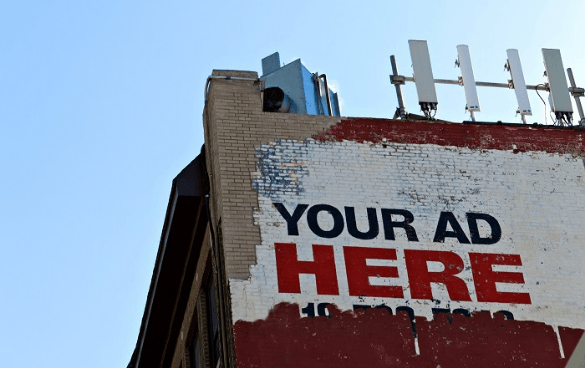 Side of a building with a faded painted advertisement stating 'YOUR AD HERE' in red letters, with a phone number below and antennas on the roof against a blue sky.