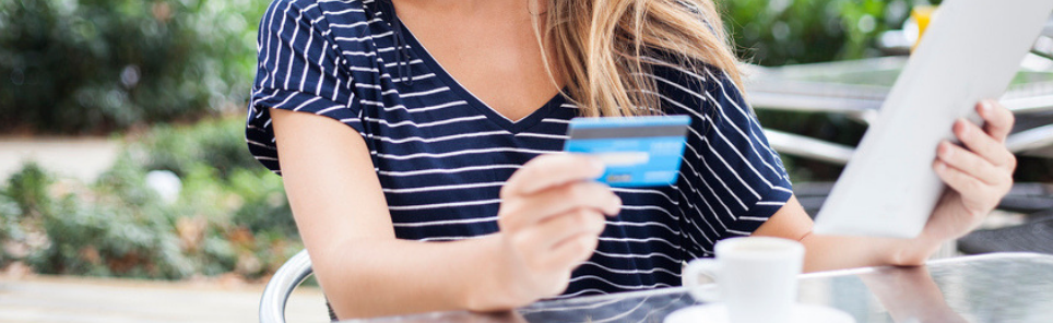 Woman holding a credit card and looking at a menu while sitting at an outdoor cafe table with a coffee cup in front of her.