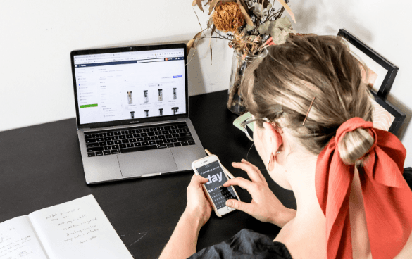 Person at a desk using a laptop with a shopping website on the screen while holding a smartphone, with a notebook and a vase with dried plants beside the laptop.