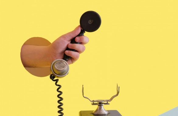 A hand holding a black telephone receiver above a vintage telephone base with ornamental horns, against a yellow background.