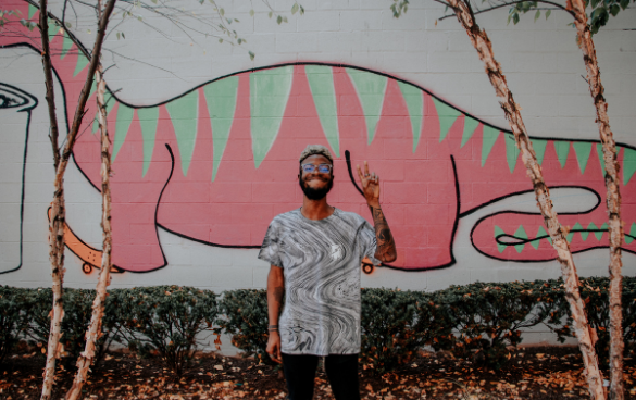 Person with glasses and a beard smiling and making a peace sign in front of a wall with a pink and green cartoonish mural, partially obscured by thin tree trunks.