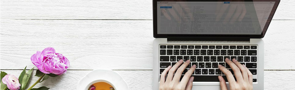 Overhead view of a person typing on a laptop next to a cup of tea and a pink peony flower on a white wooden surface.