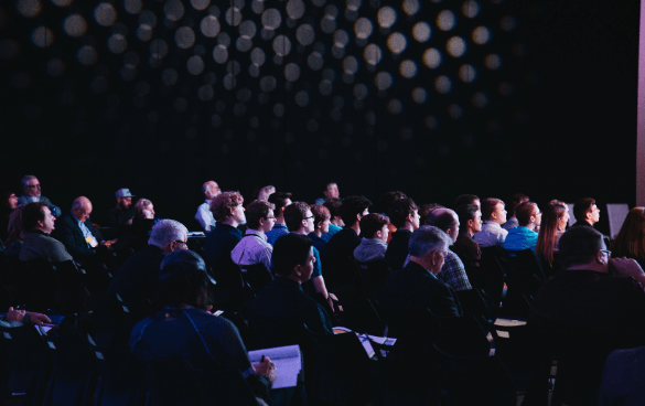 Audience members seated in rows at a dimly lit conference or seminar, attentively facing towards the front.