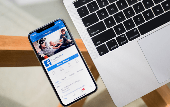 A smartphone with the Facebook profile page open, resting on a MacBook keyboard on a wooden surface.