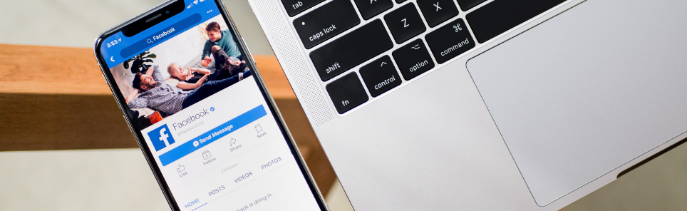 Smartphone with Facebook profile on screen next to an open laptop with visible keyboard.