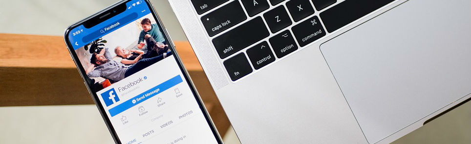 Smartphone with Facebook profile page on screen beside a laptop with black keyboard on a wooden desk.