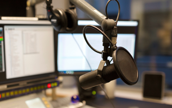 Close-up of a studio microphone with a pop filter in a radio broadcast studio with computer monitors and equipment in the background.